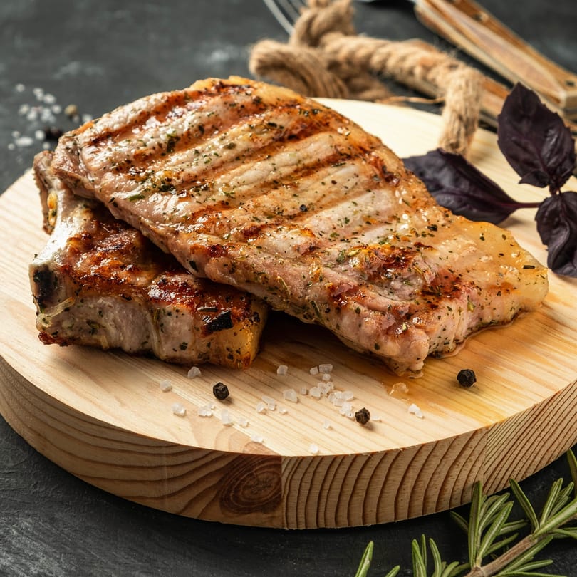 Two grilled pork chops with grill marks, seasoned with herbs, salt, and pepper on a wooden board.