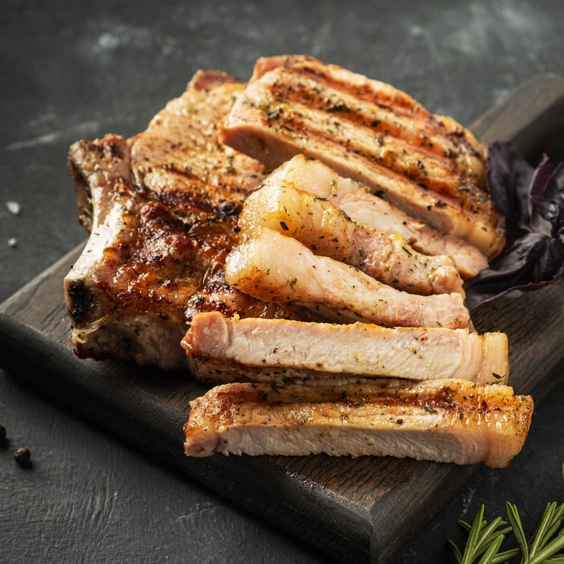 Grilled pork chops, some sliced, on a dark wooden board with herbs.